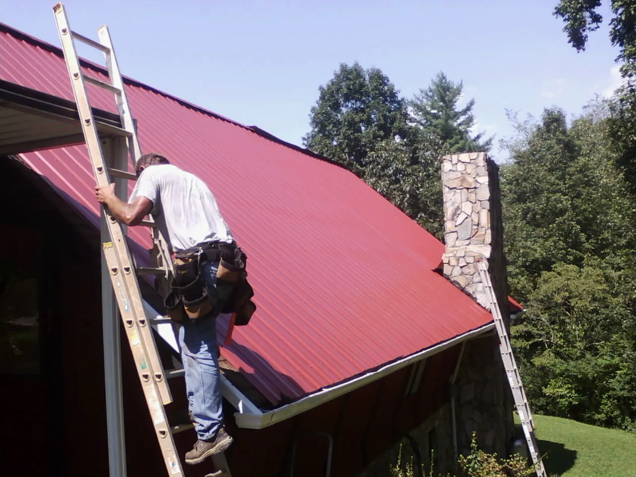 Roofer on a ladder beside a red metal roof and stone chimney — Soffit & Fascia Repair by Roofing Brothers in Coos Bay